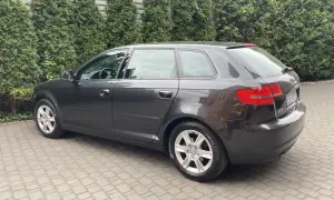 Ciemnoszary Audi A3 8P Sportback stoi na podjeździe z kostki brukowej, otoczony zielonym żywopłotem.