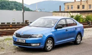 Niebieski liftback Skoda Rapid stoi na tle torów kolejowych i starych budynków.