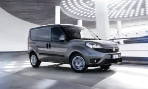 Szary Fiat Doblo Cargo w ruchu, z rozmytymi kołami, na tle nowoczesnej architektury z okrągłymi światłami.