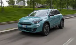 Nowoczesny, turkusowy Fiat 600 Hybrid sunie po miejskiej drodze, otoczony zielenią i budynkami.