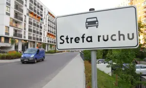 Znak "Strefa ruchu" przed nowoczesnym budynkiem mieszkalnym. Niebieski bus jedzie po drodze.