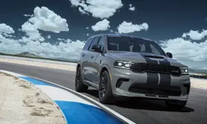 Szary Dodge Durango SRT Hellcat z czarnymi pasami pędzi po torze wyścigowym pod błękitnym niebem z chmurami.