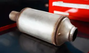 Metalowy tłumik, element układu wydechowego, leży na ciemnej powierzchni. W tle widać czerwony element, być może skrzynkę narzędziową. To część do samochodu, może nawet nowy gpf.