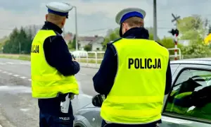 Dwóch policjantów w kamizelkach odblaskowych przy samochodzie. Jeden z nich trzyma broń. Prawdopodobnie zatrzymali kierowcę za przekroczenie prędkości o 50 km/h.
