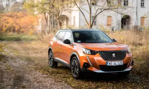 Pomarańczowy Peugeot 2008 GT na tle jesiennego krajobrazu z opuszczonym budynkiem.
