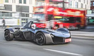 Szary BAC Mono z kierowcą w kasku pędzi ulicą, mijając rozmazany czerwony autobus piętrowy.