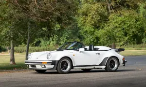 Biały Porsche 911 Speedster z lat 80. z czarnym dachem i spojlerem, zaparkowany na tle drzew.