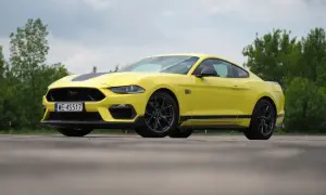 Żółty Ford Mustang Mach 1 z czarnymi pasami na masce i bokach, zaparkowany na tle zieleni.