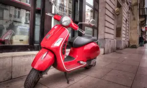 Czerwony skuter Vespa, klasyk z kategorii AM, zaparkowany na chodniku przed budynkiem z witrynami.