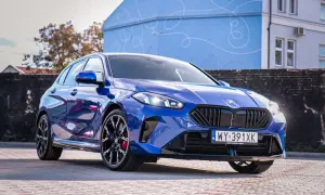 Błyszczące, niebieskie BMW 120 z czarnymi felgami stoi na brukowanym placu przed budynkiem z niebieską, ozdobną fasadą.