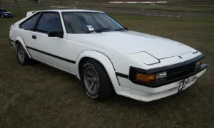 Biała Toyota Supra MK2 z lat 80. stoi na trawie, z widokiem na tor wyścigowy w tle.