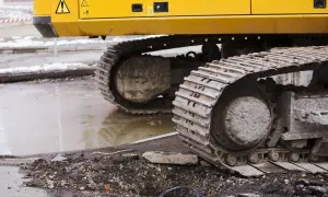 Gąsienice koparki, z widoczną zwolnicą, stoją na błotnistym terenie po pracach drogowych.