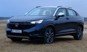 Nowa Honda HR-V w ciemnym kolorze, zaparkowana na trawiastym polu, z widokiem na jezioro i las w tle.