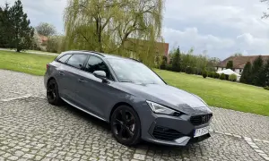 Szary Cupra kombi na brukowanym podjeździe, z zielonym trawnikiem i drzewami w tle.