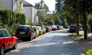 Ulica z wieloma samochodami, gdzie parkowanie na chodniku jest powszechne. Widać domy i drzewa.