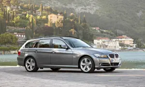 Szary kombi BMW 325i zaparkowany nad jeziorem, z malowniczymi wzgórzami i budynkami w tle.