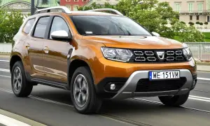 Pomarańczowy Dacia Duster 2 jedzie po moście, za nim widać budynki i drzewa.