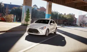 Biały Toyota Yaris Sedan jedzie pod wiaduktem, mijając budynki i drzewa.