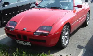 Czerwony kabriolet BMW Z1 z tablicą rejestracyjną PAX 877 zaparkowany obok czarnego samochodu.