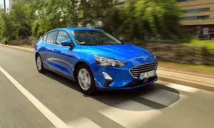 Błękitny Ford Focus sedan pędzi po miejskiej drodze, mijając drzewa i budynki.