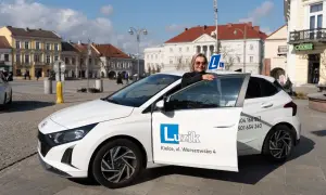 Kobieta z uśmiechem opiera się o drzwi białego samochodu szkoleniowego z literą "L" na dachu. To idealny moment, by zacząć naukę jazdy i zdobyć prawo jazdy B.