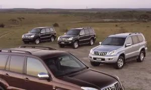 Cztery samochody Toyota Land Cruiser 120, w tym jeden brązowy, dwa czarne i jeden srebrny, stoją na polnej drodze wśród traw.