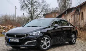 Czarny Peugeot 508 na brukowanej drodze. W tle zrujnowane budynki i drzewa. Wiele osób szuka peugeot 508 opinie przed zakupem.