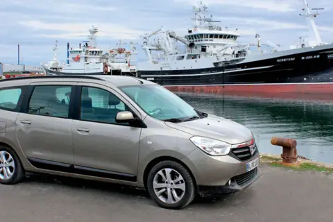 Brązowy Dacia Lodgy zaparkowany przy nabrzeżu, w tle wielkie statki rybackie.
