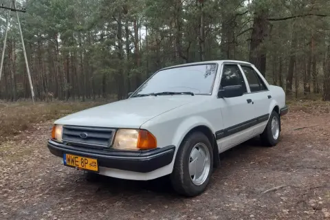 Biały Ford Orion z lat 80. stoi na leśnej drodze.