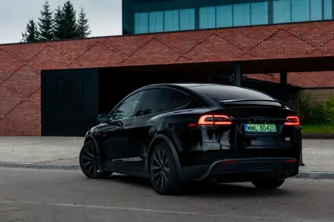 Czarna Tesla X z włączonymi światłami z tyłu, zaparkowana przed nowoczesnym budynkiem z cegły.