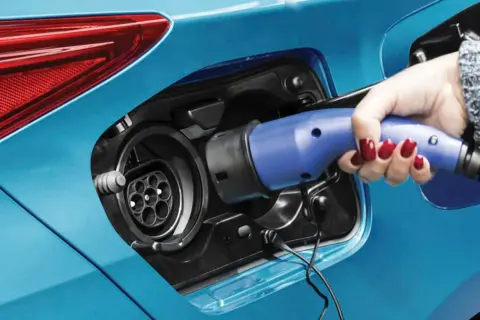 Dłoń z czerwonym lakierem na paznokciach podłącza ładowarkę do niebieskiej hybrydy plug-in.