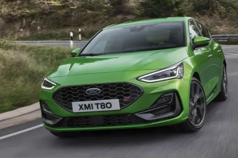 Jaskrawozielony Ford Focus 2025 ST z charakterystycznym grillem i światłami LED pokonuje zakręt na malowniczej, leśnej drodze.