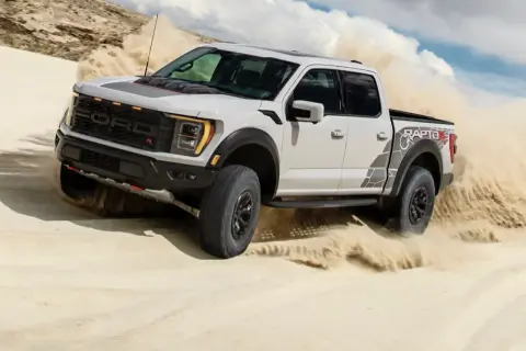 Biały Ford F-150 Raptor szaleje po piaszczystych wydmach, wzbijając tumany piasku.