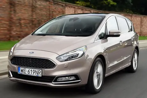 Elegancki Ford S-Max w kolorze piaskowym jedzie po drodze, z ceglanym murem w tle.