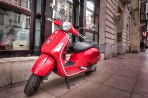 Czerwony skuter Vespa, klasyk z kategorii AM, zaparkowany na chodniku przed budynkiem z witrynami.