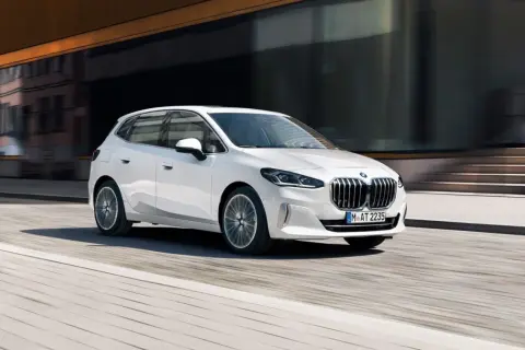Biały BMW 2 Active Tourer w ruchu miejskim, z rozmytym tłem budynków i ulicy.