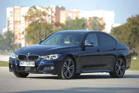 Ciemnoniebieskie BMW 318d z polskimi tablicami rejestracyjnymi stoi na drodze. W tle widać budynki i drzewa.