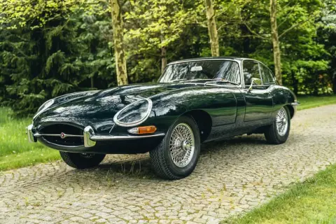 Klasyczny, ciemnozielony Jaguar E-Type na brukowanej drodze, otoczony drzewami.