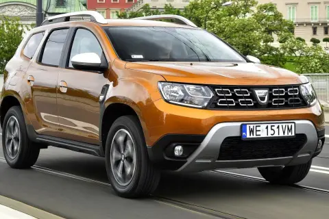 Pomarańczowy Dacia Duster 2 jedzie po moście, za nim widać budynki i drzewa.