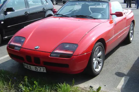 Czerwony kabriolet BMW Z1 z tablicą rejestracyjną PAX 877 zaparkowany obok czarnego samochodu.
