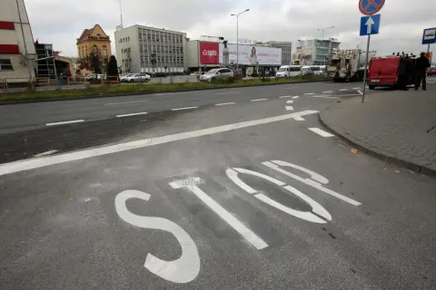 Na asfalcie widoczna jest biała linia warunkowego zatrzymania "STOP". W tle budynki i ruch uliczny.