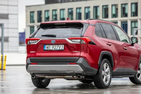 Czerwony Toyota RAV4 2020 Hybrid na mokrym parkingu, z nowoczesnym budynkiem w tle.
