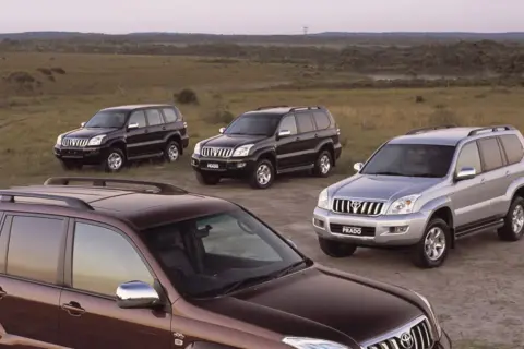 Cztery samochody Toyota Land Cruiser 120, w tym jeden brązowy, dwa czarne i jeden srebrny, stoją na polnej drodze wśród traw.