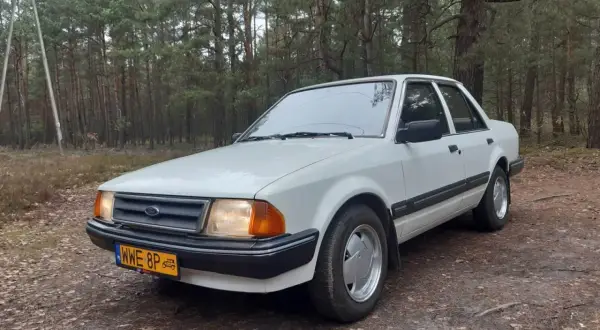 Biały Ford Orion z lat 80. stoi na leśnej drodze.