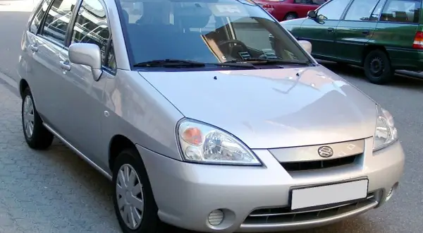 Srebrny samochód Suzuki Liana stoi na ulicy. W tle widać inne samochody i budynki.