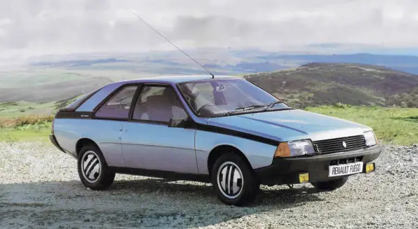 Jasnoniebieski Renault Fuego z lat 80. stoi na szutrowej drodze, z widokiem na pagórkowaty krajobraz i chmurne niebo.