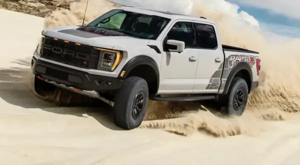 Biały Ford F-150 Raptor szaleje po piaszczystych wydmach, wzbijając tumany piasku.