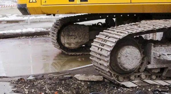 Gąsienice koparki, z widoczną zwolnicą, stoją na błotnistym terenie po pracach drogowych.