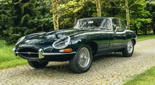 Klasyczny, ciemnozielony Jaguar E-Type na brukowanej drodze, otoczony drzewami.
