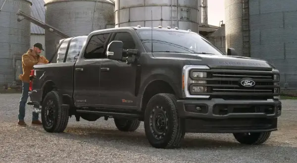 Mężczyzna ładuje pakunki do ciemnoszarego Forda Super Duty. Ciężarówka stoi na żwirowej drodze obok silosów.